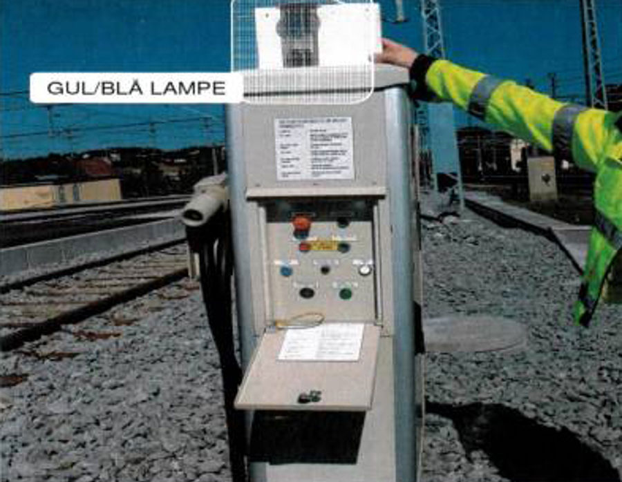 Togvarmepost med åpent betjeningspanel, knapper og lamper. Hånd peker mot gul og blå lampe øverst